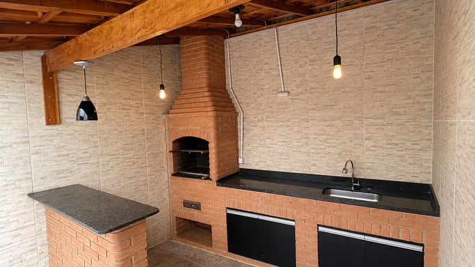 Espaço gourmet coberto com churrasqueira, bancada em granito, pia, armários inferiores, balcão americano e agradável iluminação quente.