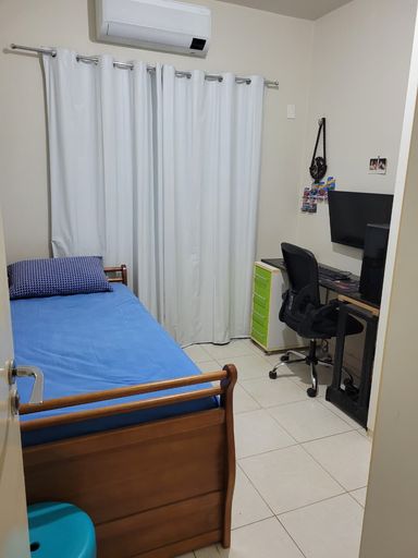 Dormitório de solteiro configurado com cama e bancada para estação de estudos/home office, apresentando piso cerâmico claro e cortina em tecido na janela no Bairro Aeroclube, em Porto Velho-RO.