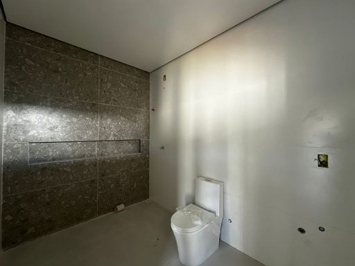 Banheiro em fase final de acabamento com bacia sanitária monobloco instalada, parede técnica revestida em porcelanato escuro texturizado com nicho embutido e teto em gesso com tabica perimetral no Bairro Triângulo, em Porto Velho-RO.