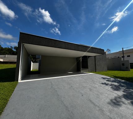 Fachada frontal contemporânea com platibanda e linhas retas, destacando a ampla garagem coberta, pintura externa em tom cinza escuro texturizado, rampas de acesso pavimentadas e áreas laterais gramadas no Bairro Triângulo, em Porto Velho-RO.