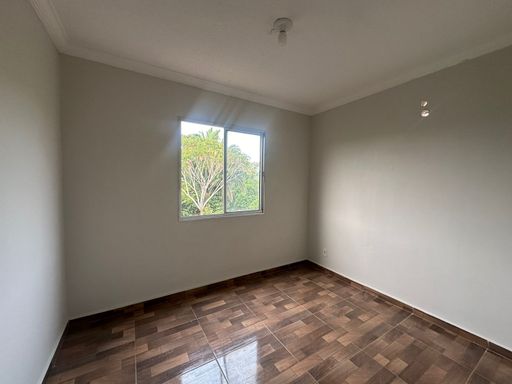 Dormitório bem iluminado com janela para área verde, garantindo ventilação natural. Com piso amadeirado e paredes claras, é um ambiente versátil em apartamento no Bairro Novo, Porto Velho/RO.
