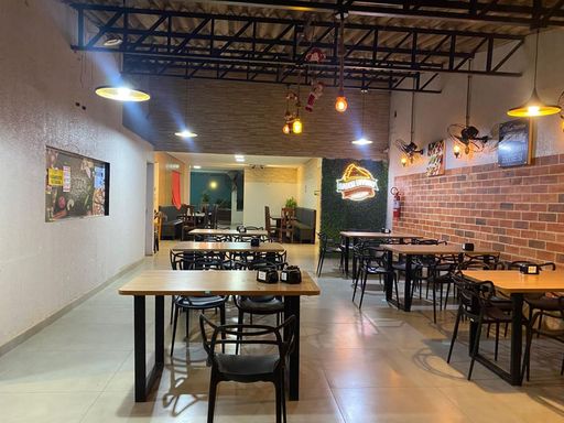 Salão principal de atendimento com piso cerâmico, teto com estrutura metálica e iluminação decorativa suspensa, contando com mesas e cadeiras distribuídas em ambiente climatizado no Bairro Esperança da Comunidade, em Porto Velho-RO.