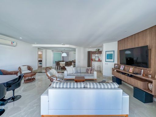 Living planejado e integrado
Amplo living com sofás, painel de madeira e mesa de jantar ao fundo.
Uma visão geral do living  mostra a disposição integrada dos móveis sob um teto rebaixado em gesso com spots de LED. Um sofá de três lugares em tom claro fica de costas para o observador, direcionado ao painel de TV em madeira que ocupa a parede lateral. Ao fundo, avista-se a sala de jantar e outras áreas de convivência. O uso de tecidos leves nos estofados e o piso polido conferem frescor e uma estética contemporânea e clean ao imóvel.
