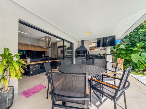 Sala de jantar e área gourmet

Mesa de jantar integrada à área gourmet com churrasqueira.

Esta sala de jantar sofisticada apresenta uma grande mesa de madeira maciça com tampo em mármore creme, acompanhada por dez cadeiras estofadas em linho bege. O ambiente é integrado à área gourmet por meio de portas de correr de vidro, que revelam uma churrasqueira embutida em parede revestida de pedra. O piso é em porcelanato claro, e as paredes são brancas. A iluminação é suave, com pontos de luz embutidos no teto e um pendente central sobre a mesa. O ambiente transmite elegância e conforto.