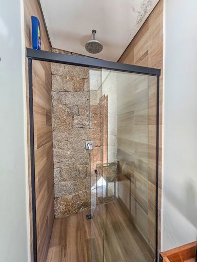 Área de banho com ducha

Box de vidro integrado a parede de pedra natural e revestimento amadeirado.

O espaço de ducha apresenta um box de vidro com estrutura preta, emoldurando uma parede lateral de pedra natural bruta. As demais paredes utilizam revestimento cerâmico que imita madeira clara, criando um contraste acolhedor com o mineral frio. Uma ducha de teto moderna garante funcionalidade e luxo. A estética é de um spa moderno e integrado a elementos naturais, transmitindo uma sensação térmica de relaxamento imediato e higiene impecável.