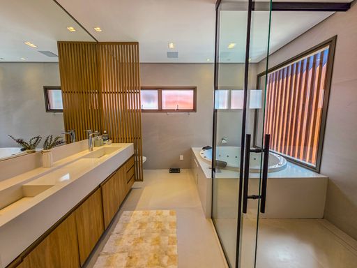 Banheiro master com hidromassagem

Área de SPA privativa com bancada de mármore, pia dupla e banheira redonda.

O banheiro principal é um verdadeiro spa, equipado com uma generosa banheira de hidromassagem circular sob janelas com brises de madeira para privacidade. A bancada longa em mármore claro possui cubas duplas e amplo espelho superior. Um painel ripado de madeira separa a zona de banho da área seca, aquecendo a estética do revestimento cerâmico bege. A sensação térmica é de relaxamento e luxo, com ventilação natural abundante e iluminação zenital suave.