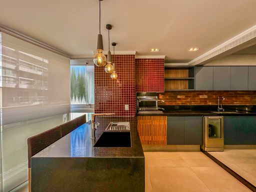 Área gourmet com churrasqueira.

Bancada de granito preto com pia e churrasqueira embutida em parede de azulejos vermelhos.

Esta área gourmet privativa é revestida com azulejos quadrados em tom vermelho escuro, criando um ponto focal vibrante. A bancada em granito preto inclui uma cuba em inox e espaço para preparo de carnes ao lado da churrasqueira. Pendentes modernos de filamento garantem uma iluminação quente e acolhedora sobre o balcão. O ambiente é planejado para recepções sociais, unindo a praticidade da pedra com a estética marcante dos revestimentos cerâmicos.