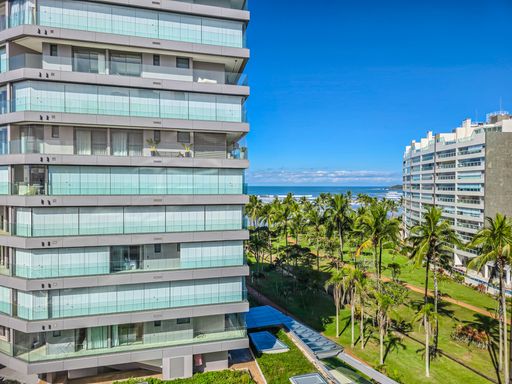 Detalhe da fachada e jardins.

Vista lateral de edifício residencial com jardim tropical e mar ao fundo.

Esta fotografia foca na relação entre o edifício e sua área verde privativa. A estrutura vertical com sacadas de vidro reflete o azul do céu, enquanto o térreo é adornado por coqueiros e um gramado bem cuidado. Ao horizonte, é possível avistar a linha do mar, confirmando a localização privilegiada do imóvel. Materiais como vidro, metal e alvenaria clara dominam a cena, proporcionando uma estética limpa e arejada, típica de empreendimentos de luxo na costa.