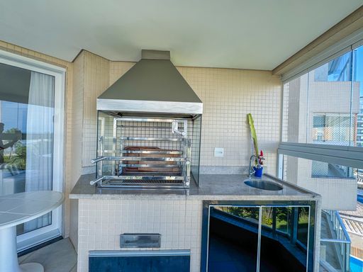 Churrasqueira moderna em aço inox
Detalhe em close de uma churrasqueira gourmet de inox com coifa em bancada de granito.
Visão detalhada da área gourmet com churrasqueira de alto padrão fabricada em aço inoxidável e equipada com espetos giratórios. A estrutura está perfeitamente embutida em uma bancada de granito cinza escuro polido, que oferece suporte funcional e pia integrada. A parede ao fundo apresenta revestimento cerâmico texturizado em tons neutros, ressaltando o design moderno e a alta durabilidade dos materiais, perfeitos para um ambiente náutico e litorâneo sofisticado.