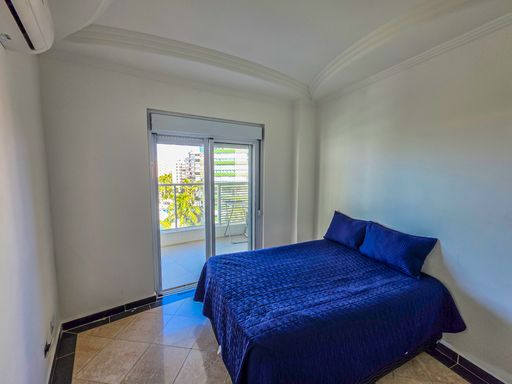 Dormitório de casal em azul.

Quarto com cama de casal azul e vista para o exterior.

O dormitório principal exibe uma cama de casal com colcha em azul marinho vibrante, contrastando com as paredes brancas. Uma ampla janela com esquadrias de alumínio oferece vista para a área externa arborizada, garantindo ventilação natural. O piso cerâmico em tons pastéis e o acabamento em gesso no teto conferem sofisticação. A iluminação é suave, ideal para um ambiente íntimo. A estética é leve e relaxante, com uma sensação térmica fresca devido à posição privilegiada no edifício.