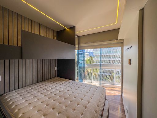 Dormitório com sancas iluminadas

Quarto com cama de casal e iluminação de LED embutida no teto.

Este dormitório apresenta um projeto de iluminação moderno, com sancas de LED de tom amarelado que percorrem o perímetro do teto. A parede da cabeceira conta com um painel cinza escuro e revestimento texturizado que remete a tecidos finos. Uma ampla janela lateral permite a entrada de luz natural e oferece vista para a sacada envidraçada. O piso laminado de madeira traz conforto térmico à área íntima, enquanto a paleta de cores neutras proporciona uma estética de tranquilidade e sofisticação para o repouso.