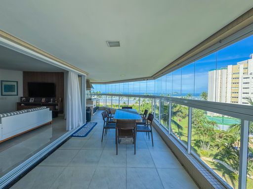 Varanda gourmet ampla com vista
Varanda de apartamento com fechamento em vidro, mesa de jantar e vista para área verde e o mar.
Esta espaçosa varanda gourmet de alto padrão destaca-se pelo fechamento panorâmico em cortina de vidro, que proporciona excelente iluminação natural e uma deslumbrante vista mar. O ambiente conta com piso em porcelanato claro e uma grande mesa retangular com tampo de pedra escura, cercada por cadeiras de fibra sintética marrom. A integração com o living interno cria uma sensação de amplitude, sofisticação e frescor praiano, ideal para momentos de lazer e descanso.