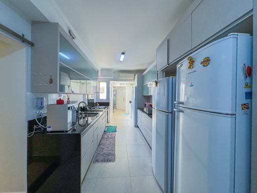 Cozinha funcional com geladeira duplex
Cozinha planejada em corredor destacando geladeira branca e bancadas escuras.
Esta tomada da cozinha em image_65a006.jpg destaca, em primeiro plano à direita, uma geladeira duplex branca decorada com pequenos ímãs. As bancadas paralelas de granito preto polido criam uma superfície contínua para eletrodomésticos e preparo de alimentos, contrastando com os móveis planejados superiores de tom azulado. A iluminação direta e o formato retilíneo garantem um excelente aproveitamento de espaço, mantendo o ambiente prático e moderno para o dia a dia.
