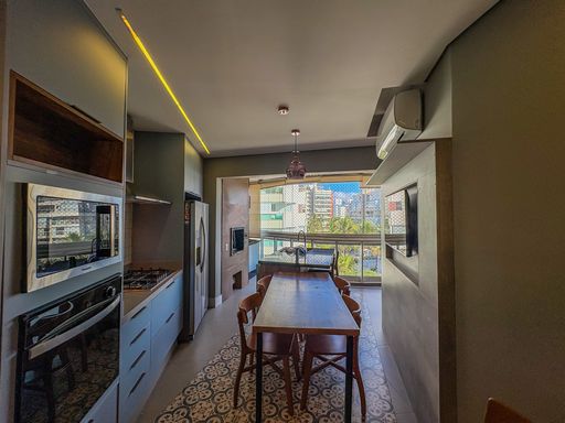 Cozinha gourmet integrada

Área de jantar e cozinha com bancada de granito e sancas de LED.

Este ambiente de conceito aberto integra a cozinha planejada à sala de jantar através de uma extensa mesa de madeira maciça. A cozinha conta com bancadas em granito preto, armários em tons de cinza azulado e eletrodomésticos em aço inox embutidos. O teto de gesso exibe sancas de LED que percorrem todo o comprimento da área gourmet, criando uma iluminação funcional e elegante. A vista da sacada envidraçada ao fundo garante luz natural, transmitindo uma estética de integração social e modernidade culinária.