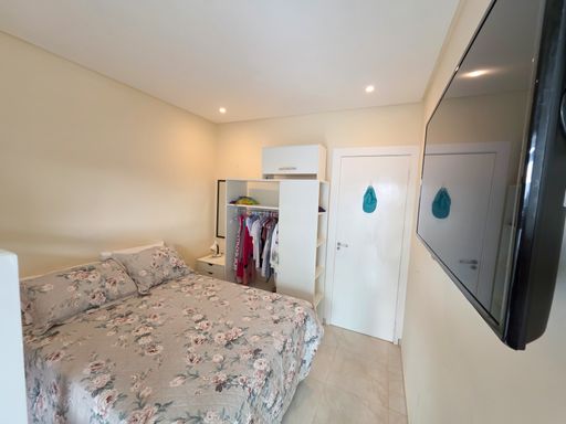 Quarto aconchegante com guarda-roupa

Cama de casal com estampa floral e guarda-roupa compacto com espelho.

Perspectiva interna do quarto destacando a organização do mobiliário. A cama de casal com enxoval floral cinza e rosa ocupa a lateral do cômodo, posicionada em frente a uma parede equipada com uma televisão de tela plana embutida. Ao fundo, um guarda-roupa modular branco com portas abertas revela um cabideiro organizado e uma gaveta espelhada. A porta de passagem branca possui um adorno decorativo azul, e o teto rebaixado em gesso com spots de iluminação embutidos cria um clima intimista, relaxante e muito acolhedor.Quarto iluminado com acesso externo

Cama de casal com colcha floral posicionada próxima a porta de correr de vidro.

Um quarto aconchegante com piso cerâmico claro e paredes pintadas em tom suave de amarelo-creme. O mobiliário inclui uma cama de casal vestida com enxoval de estampa floral e uma cadeira de madeira com assento azul disposta ao lado. Uma grande porta de correr de vidro com esquadria branca se abre para o terraço externo, inundando o ambiente com luz natural. Um aparelho de ar-condicionado split instalado no alto garante o conforto térmico, complementado por um mancebo de chão para pendurar pertences e roupas.