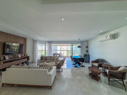 Living amplo voltado para varanda
Sala de estar integrada com sofás claros, ar-condicionado e vista para varanda.
A perspectiva ampliada da área social  destaca a conexão fluida entre a sala de estar e a varanda gourmet ao fundo. Um sofá branco com almofadas listradas compõe o centro do espaço, que é guarnecido por móveis de vime escuro nas laterais. Na parede à direita, um aparelho de ar-condicionado garante o conforto térmico, complementando o ambiente clean iluminado pela claridade natural do dia que reflete no piso cerâmico brilhante.