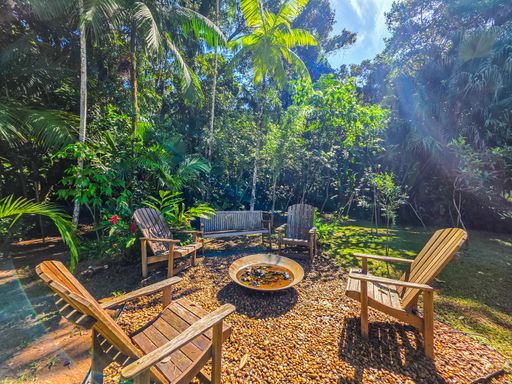 Área de fogueira externa

Espaço lounge de jardim com cadeiras de madeira ao redor de lareira externa.

Este espaço de convivência noturna ou de inverno dispõe de cadeiras de madeira rústica dispostas circularmente em torno de um "fire pit" central sobre pedriscos. Cercado por vegetação densa e árvores, o ambiente oferece privacidade e uma conexão direta com a natureza. A estética é acolhedora e "boho", ideal para reuniões sociais informais ao ar livre. A sensação térmica remete ao conforto de uma fogueira sob a copa das árvores, integrando rústico e moderno.