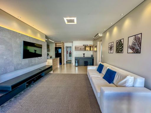 Living contemporâneo com iluminação LED.

Sala ampla com sofá branco, tapete cinza e painel de TV iluminado.

Uma perspectiva ampla do living que evidencia o design minimalista e a funcionalidade do espaço. O teto rebaixado em gesso conta com fitas de LED embutidas e spots direcionais que realçam as texturas das paredes e dos quadros decorativos. O tapete de fibra natural em tom cinza delimita a área de convivência, harmonizando com o piso frio e a marcenaria de alto padrão. A paleta de cores neutras é pontuada pelo azul vibrante das almofadas, transmitindo uma estética de serenidade e conforto térmico, ideal para momentos de relaxamento em um imóvel de alto padrão.