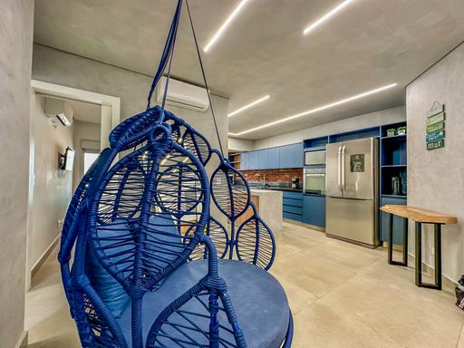 Vista da sala para cozinha.

Poltrona suspensa azul em primeiro plano com visão da cozinha integrada ao fundo.

A fotografia demonstra a fluidez do conceito aberto, conectando a sala de estar à cozinha planejada. A poltrona suspensa de design vazado azul domina o primeiro plano, enquanto ao fundo observa-se a bancada da cozinha em tons de azul e madeira. A iluminação linear do teto percorre os dois ambientes, unificando o projeto arquitetônico. Materiais como fibra, metal e madeira se misturam, criando uma estética moderna e dinâmica. A sensação é de um imóvel bem ventilado e com excelente aproveitamento de espaço.