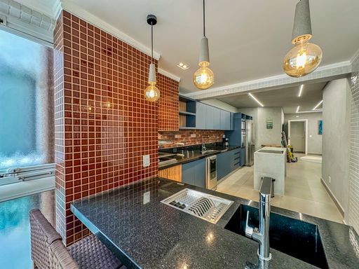 Detalhe da área gourmet.

Bancada de granito preto com cuba e torneira cromada sob luzes pendentes.

Visão aproximada da bancada gourmet, destacando o acabamento polido da pedra negra e a cuba profunda em aço inox. Três luminárias pendentes com lâmpadas de estilo industrial descem do teto, proporcionando luz direta sobre a área de trabalho. Ao fundo, os azulejos vermelhos e os armários escuros completam a paleta de cores sofisticada. O ambiente transmite uma sensação de hospitalidade e bom gosto, sendo um espaço chave para o lazer em apartamentos modernos.