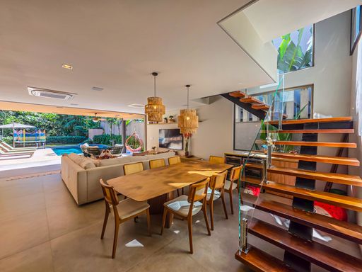 Ambiente interno com mesa de jantar longa em madeira e escada lateral.  

Este amplo espaço de convivência apresenta uma mesa de jantar robusta feita de madeira maciça, acompanhada por cadeiras de design clássico. À direita, uma escada de madeira com estrutura metálica preta e guarda-corpo de vidro confere um toque industrial sofisticado. O teto branco possui iluminação embutida e pendentes de fibra natural que trazem uma textura orgânica ao ambiente. A integração total com a área externa através de portas de vidro garante luminosidade natural e uma sensação térmica agradável e arejada.