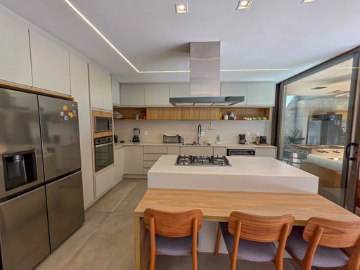 Ilha de cozinha gourmet

Bancada branca com cooktop integrado e mesa de refeições em madeira anexa.

A ilha central funciona como ponto de cocção com cooktop embutido e coifa cilíndrica de inox acima. Anexa à ilha, uma mesa de madeira clara com banquetas modernas permite refeições rápidas e interação constante. Ao fundo, as paredes são ocupadas por armários que abrigam fornos e geladeira de forma discreta. A iluminação por fita LED embutida no gesso acentua as linhas retas do mobiliário, criando uma estética contemporânea e tecnologicamente eficiente.