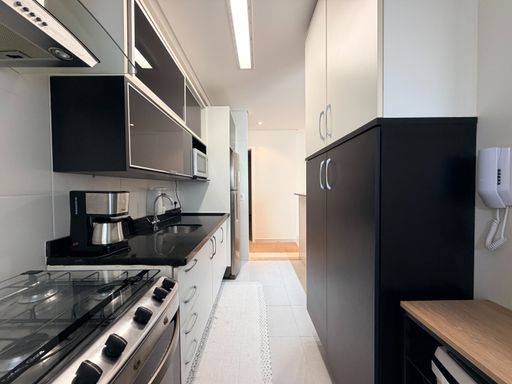 Cozinha planejada funcional e corredor
Cozinha em corredor com armários pretos e brancos e bancada escura.
A visão interna da cozinha detalha o excelente aproveitamento de espaço no formato corredor. A bancada comprida de granito preto polido acomoda uma pia de inox e uma cafeteira elétrica, posicionada ao lado do fogão embutido. O contraste elegante entre os armários aéreos pretos e os gabinetes brancos é complementado por uma torre de armários escuros à direita. A iluminação em calha embutida no teto garante clareza e uma estética limpa.