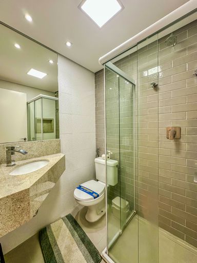 Banheiro social com bancada de granito

Banheiro com bancada em granito claro, pia embutida, espelho amplo e box de vidro com revestimento cerâmico cinza.

Este banheiro social bem distribuído possui uma bancada funcional de granito ornamental em tons de bege e marrom, com cuba embutida oval branca e torneira de duplo comando cromada. Um generoso espelho plano cobre a parede acima da bancada, ampliando visualmente o espaço. A área do box é isolada por portas de vidro temperado transparente e conta com paredes revestidas em cerâmica retangular cinza escuro brilhante, oferecendo um contraste moderno com as texturas claras e laváveis do restante do cômodo.