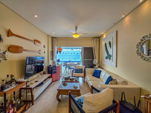 Sala de TV com vista externa

Sala de estar decorada com sofás e vista para a varanda.

Perspectiva inversa da sala de estar, enfatizando a transição para a varanda gourmet por meio de uma grande porta de correr de vidro. O espaço é composto por um sofá claro com detalhes em azul, poltronas de madeira e fibra trançada e uma mesa de centro com objetos decorativos. A iluminação aconchegante provém de spots direcionais no teto e do ventilador com luminária. Elementos decorativos nas paredes, como quadros de folhas verdes e os remos de madeira, reforçam a sensação estética praiana e sofisticada do imóvel.