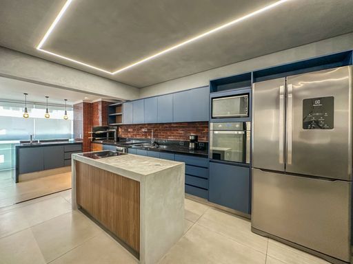 Cozinha planejada azul moderna.

Cozinha americana com armários azuis, bancada de granito preto e ilha central de cimento.

Esta cozinha planejada de alto padrão combina armários em azul fosco com bancadas de granito preto e uma ilha central robusta com acabamento em cimento e madeira. A iluminação linear no teto garante visibilidade perfeita sobre as áreas de trabalho, que incluem cooktop e forno embutidos. O revestimento de tijolinhos aparentes atrás da pia adiciona uma textura rústica que aquece a estética industrial predominante. A disposição é funcional e ideal para entusiastas da gastronomia, transmitindo uma sensação de modernidade e eficiência.