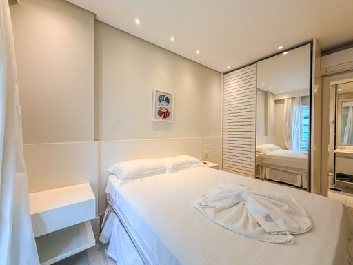Quarto de casal com guarda-roupa espelhado

Quarto de casal exibindo guarda-roupa com portas de espelho amplas e cama de casal.

Visão lateral do quarto de casal com foco no grande guarda-roupa planejado que possui portas de correr totalmente revestidas de espelhos, duplicando visualmente a percepção de espaço e refletindo a luz natural proveniente da varanda. A cama de casal está arrumada com lençóis claros e colcha texturizada, ladeada por um criado-mudo branco minimalista com uma gaveta. A iluminação indireta feita por spots embutidos no teto de gesso destaca a paleta de cores suaves (bege, off-white e branco) e a parede texturizada da cabeceira, resultando em um espaço sofisticado, luxuoso e relaxante.