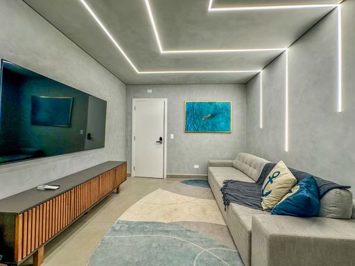 Living integrado com TV.

Painel de TV em cimento queimado com rack ripado e iluminação linear.

A perspectiva foca na área de entretenimento, destacando o painel de TV integrado à parede de cimento queimado. O rack inferior apresenta acabamento em madeira ripada, conferindo textura e elegância ao ambiente. No teto, as linhas de luz LED criam um desenho geométrico que guia o olhar pelo espaço. Um tapete com formas orgânicas e tons de azul e cinza complementa a disposição dos móveis, enquanto um quadro abstrato de oceano reforça a estética litorânea moderna. A sensação térmica é de frescor e amplitude.