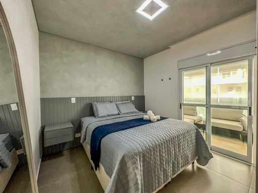 Suíte máster moderna.

Quarto de casal com cama box cinza, painel ripado e ar-condicionado.

O dormitório principal segue a estética industrial com paredes de cimento queimado e um painel de cabeceira em madeira cinza ripada. A cama box está vestida com enxoval cinza e uma peseira azul marinho, mantendo a paleta de cores do imóvel. Um ar-condicionado Split garante o conforto térmico, enquanto as janelas de correr permitem a entrada de luz natural lateral. A iluminação de teto é feita por um plafon quadrado moderno, criando uma sensação de refúgio urbano sereno.