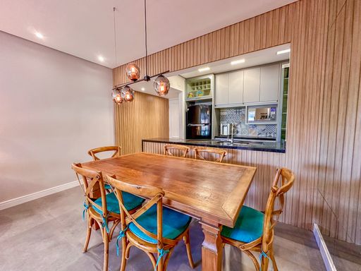 Sala de jantar integrada à cozinha

Mesa de jantar de madeira e cadeiras azuis integradas à bancada americana.

Esta perspectiva realça o conceito aberto de convivência unindo a sala de jantar, a cozinha planejada e a varanda ao fundo. A mesa de madeira com cadeiras azuis está disposta estrategicamente ao lado de um painel ripado vertical que camufla os acessos internos. Luminárias de design geométrico projetam uma luz suave sobre os móveis amadeirados. O revestimento cerâmico de tons neutros cinzas nas paredes contribui para uma estética limpa, contemporânea e de alto padrão imobiliário, mantendo o ambiente arejado e muito convidativo.