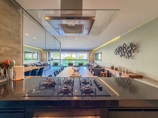 Cozinha com ilha e cooktop

Bancada de cozinha com cooktop em destaque e vista para o living.

A perspectiva da cozinha a partir da ilha central mostra o cooktop embutido na bancada de granito preto, com uma coifa de inox e vidro acima. O ambiente é integrado ao social por meio de uma parede de vidro, permitindo a comunicação visual entre quem cozinha e os convidados na sala de jantar. Os materiais nobres e a iluminação embutida reforçam o caráter gourmet e moderno da residência.