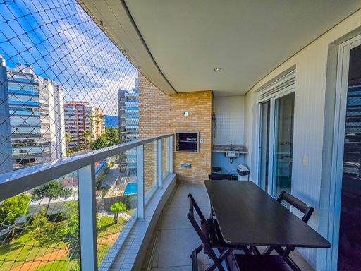 Varanda gourmet em perspectiva oposta.

Vista da varanda em direção ao interior do apartamento.

Nesta foto, observa-se a transição da varanda gourmet para o interior através de grandes portas de vidro de correr com esquadrias brancas. A churrasqueira lateral com bancada de apoio em granito destaca-se pela praticidade. A parede externa em tijolinhos claros harmoniza com o restante da fachada do edifício. A iluminação natural é o ponto forte, criando um ambiente vibrante e solar que remete à hospitalidade e ao lazer típico de residências de veraneio.