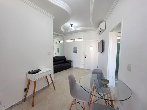 Visão geral da área social.

Sala integrada vista do canto com mesa de vidro.

Uma perspectiva ampla da área social que revela a integração inteligente entre a sala de estar, jantar e o acesso aos dormitórios. O uso de espelhos na parede lateral duplica visualmente o espaço, refletindo a luz e a decoração minimalista. O ambiente é climatizado com ar-condicionado Split, garantindo conforto térmico constante. A disposição dos móveis favorece a circulação, enquanto a iluminação em trilhos e spots pretos adiciona um toque industrial moderno à estética predominantemente clara e elegante.