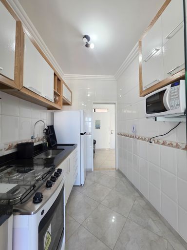 Cozinha compacta em plano aberto.

Corredor de cozinha com fogão, micro-ondas e pia.

Uma visão detalhada da cozinha mostrando a disposição linear dos equipamentos, como o fogão embutido e o micro-ondas suspenso para economizar espaço de bancada. O acabamento em aço inox de alguns utensílios e os puxadores metálicos dos armários adicionam um toque de modernidade. A janela ao fundo garante ventilação natural e iluminação durante o dia, mantendo o ambiente arejado e agradável para o preparo de refeições. A sensação é de um espaço prático e bem aproveitado.