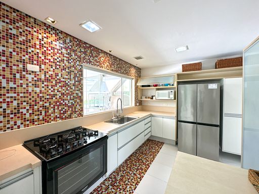 Cozinha planejada com pastilhas coloridas

Cozinha moderna com armários brancos, bancada bege, geladeira de inox e parede revestida de pastilhas vermelhas e amarelas.

Esta cozinha de design contemporâneo destaca-se pela parede de fundo inteiramente revestida por pastilhas de vidro em tons quentes de vermelho, laranja e amarelo. A bancada em quartzo bege claro abriga uma cuba embutida em aço inox com torneira monocomando e um cooktop preto de cinco bocas integrado ao forno. Os armários planejados inferiores possuem acabamento branco com puxadores em perfil de alumínio discreto. À direita, uma grande geladeira side-by-side em aço inoxidável completa o espaço. O piso em porcelanato branco e a iluminação embutida no gesso garantem um ambiente claro, higiênico e vibrante.