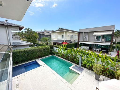 Vista aérea da área de lazer

Visão panorâmica superior da piscina, hidromassagem e jardim de condomínio fechado.

Uma perspectiva elevada revela o layout completo do quintal, mostrando a piscina principal integrada a uma área menor de hidromassagem quadrada. O deck de pedra clara contrasta lindamente com a água esverdeada e o paisagismo tropical composto por plantas de folhas largas e uma cerca viva perfeitamente aparada. Ao fundo, observam-se as fachadas de outras residências de alto padrão do condomínio sob um céu azul claro. A imagem transmite uma estética de exclusividade, amplitude e tranquilidade urbana de veraneio.