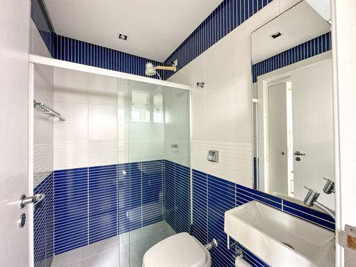 Banheiro moderno azul e branco

Banheiro com paredes revestidas em pastilhas azuis e brancas, box de vidro e pia.

Este banheiro social apresenta uma paginação marcante composta por pastilhas de vidro azul-cobalto na metade inferior e superior das paredes, divididas por uma faixa central de azulejos brancos horizontais. O ambiente possui um box de canto com portas de correr em vidro temperado transparente e esquadrias de alumínio. A cuba de apoio branca retangular está instalada sobre uma bancada suspensa compacta, acompanhada por um espelho vertical que vai até o teto. A iluminação embutida realça as cores frias, conferindo um aspecto limpo, refrescante e jovem ao espaço.