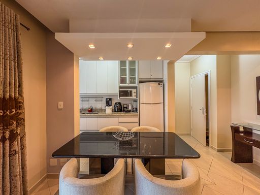 Mesa de jantar e cozinha planejada

Mesa de jantar de granito escuro em frente a cozinha americana equipada.

Visão frontal da área de refeições destacando o design contemporâneo do mobiliário. A mesa de jantar com tampo de pedra escura polida contrasta com o revestimento claro das cadeiras estofadas e texturizadas. Logo acima, uma sanca estrutural de gesso abriga spots embutidos que direcionam a luz sobre a mesa. Ao fundo, a cozinha americana revela-se compacta e equipada, apresentando geladeira duplex branca, forno de micro-ondas embutido em nicho e armários planejados que otimizam o espaço vertical de maneira organizada e limpa.