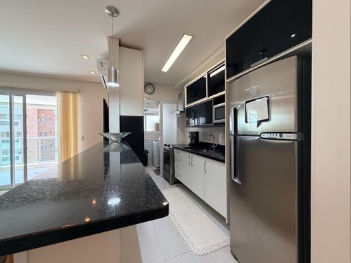 Cozinha americana com balcão gourmet
Cozinha planejada com balcão de granito preto e geladeira em aço inox.
Esta moderna cozinha em estilo americano é marcada por um generoso balcão de granito preto absoluto que divide os ambientes. O espaço é equipado com armários planejados aéreos pretos com perfis de alumínio e armários inferiores brancos. Uma grande geladeira duplex em aço escovado e uma luminária pendente cilíndrica sobre o balcão reforçam a estética contemporânea e funcional, transmitindo organização e alta praticidade.