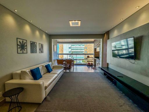 Sala de estar moderna integrada.

Sala de estar com sofá branco, painel de TV e iluminação embutida.

Esta sala de estar de conceito aberto apresenta uma estética contemporânea e sofisticada, destacando-se pelo amplo sofá em tecido branco decorado com almofadas azuis. O ambiente é revestido com piso de porcelanato neutro e possui um painel de TV em marcenaria escura que contrasta com as paredes claras. A iluminação é um ponto forte, utilizando um lustre central quadrado e diversos spots de LED embutidos no teto de gesso, criando uma atmosfera acolhedora e bem distribuída. A disposição dos móveis favorece a circulação e a integração com a varanda ao fundo, proporcionando uma sensação térmica arejada e luxuosa.
