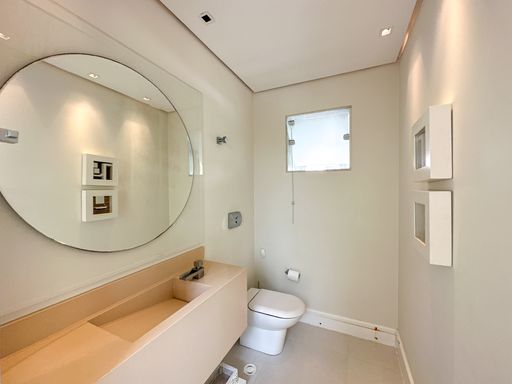 Lavabo moderno com espelho redondo

Lavabo clean com bancada esculpida bege, espelho circular e nichos iluminados.

Este sofisticado lavabo aposta em uma paleta de cores neutras e suaves para transmitir elegância. Conta com uma cuba esculpida na própria pedra de quartzo bege com torneira metálica de bica baixa. Um grande espelho perfeitamente circular domina a parede principal, refletindo a iluminação embutida acolhedora do teto de gesso. Dois nichos quadrados com moldura branca na parede lateral oferecem espaço para pequenos objetos decorativos. O vaso sanitário branco convencional harmoniza-se com o estilo minimalista e contemporâneo do espaço.