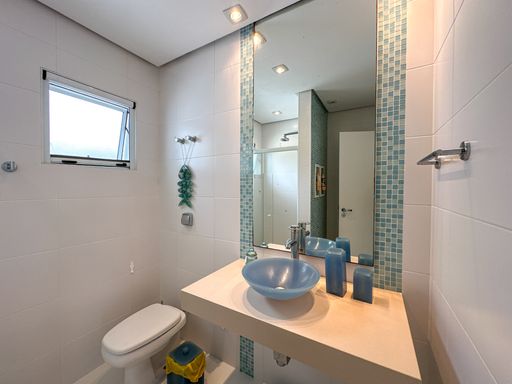 Banheiro com cuba azul decorado

Banheiro com cuba de vidro azul redonda, espelho com pastilhas e nichos.

Este banheiro compacto combina funcionalidade e design litorâneo. Possui uma bancada suspensa de quartzo bege claro com uma cuba de apoio redonda em vidro azul translúcido. O grande espelho retangular é emoldurado nas laterais por faixas verticais de pastilhas de vidro em tons de azul e verde-água. A parede oposta conta com um vaso sanitário branco e uma pequena janela basculante de alumínio que garante a ventilação do espaço. Elementos decorativos marinhos suspensos adicionam um charme extra ao ambiente clean e refrescante.