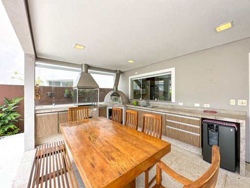 Mesa gourmet e bancada planejada

Mesa de madeira em destaque na varanda integrada com armários planejados e churrasqueira.

Uma perspectiva mais próxima da área de refeições gourmet exibe o tampo polido da mesa de madeira de demolição. Ao fundo, a bancada funcional revela armários embutidos com revestimento amadeirado sob a pia e uma chopeira ou frigobar embutido na cor preta. As grandes coifas de aço inoxidável da churrasqueira e do forno dominam a parede de fundo clara. O espaço é aberto nas laterais, garantindo uma excelente circulação de ar, frescor térmico e integração visual constante com o jardim externo da residência.