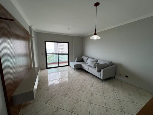 Sala de dois ambientes com painel de TV, sofá cinza com sacada com vista para o mar.