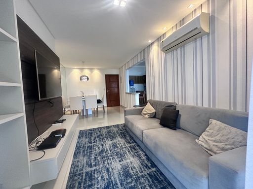 Sala ampla com dois ambientes, com ar-condicionado, sofá, mesa de jantar, parede espelhada e um elegante lustre pendente.