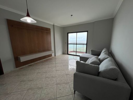Sala de dois ambientes com painel de TV, sofá cinza com sacada com vista para o mar.